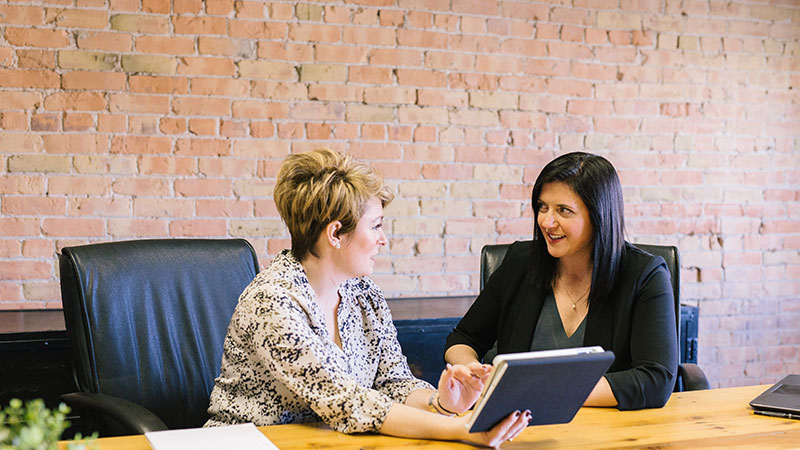 Two women sitting in a conference room having a conversation while one gestures to a book in her hand