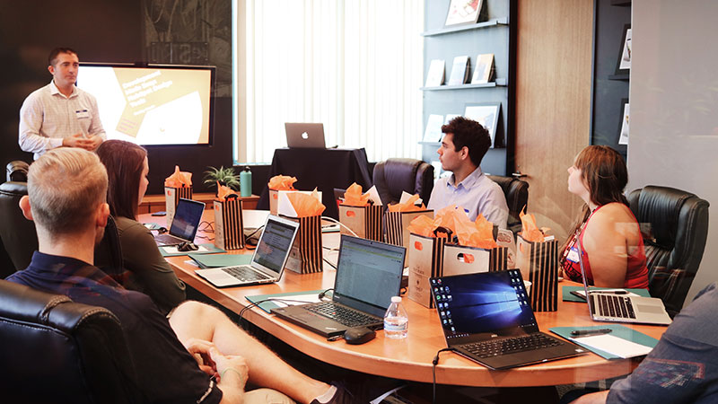 A man giving a presentation to his coworkers in a conference room. There is a table in the middle of the room with laptops and gift bags in front of each person sitting at the table.